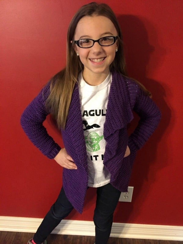 Purple example of the child size of the Blanket Cardigan crochet pattern being worn by a smiling female child.
