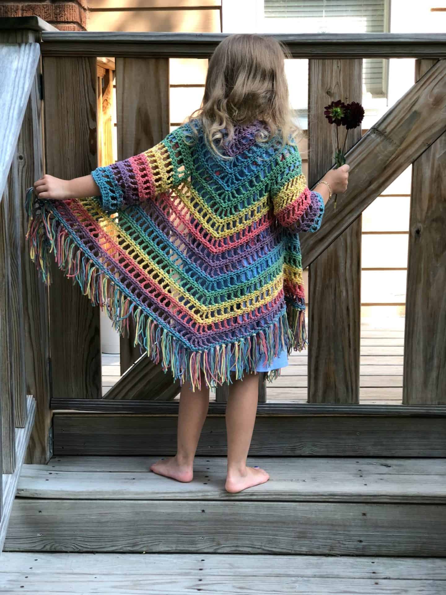 Female child wearing a colorful example of the Child Size Shawl Good Cardigan crochet pattern while standing barefoot on a wood deck.