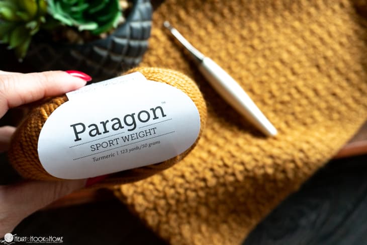 Sample of a Turmeric colorway of the Crochet Cluster Cowl pattern along with a Furls crochet hook and a skein of Turmeric colored Paragon Sport yarn.