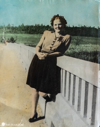Old picture of a smiling woman wearing a buttoned up shirt and skirt leaning on a rail of a bridge.