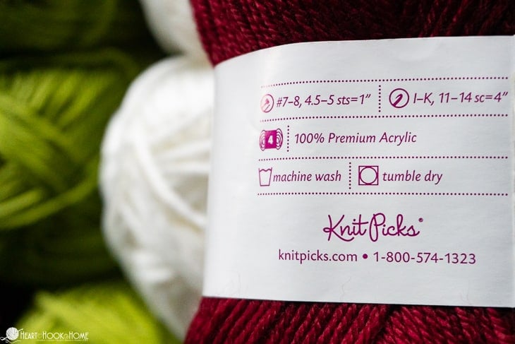 Wine colored Brava Worsted weight yarn label with white and green colored skeins in the background.