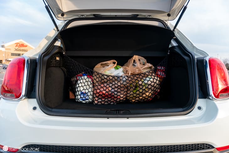 Groceries in bags being secured by an example of a black Trunk Cargo Net crochet pattern inside of a white vehicle with its trunk raised. 