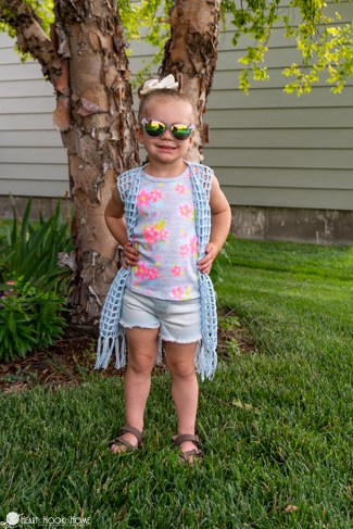 Example of the Child Size Summer Vest crochet pattern in a light blue colorway on a smiling female child wearing a flowery top and white shorts in a yard with a tree, flowers, and grass.