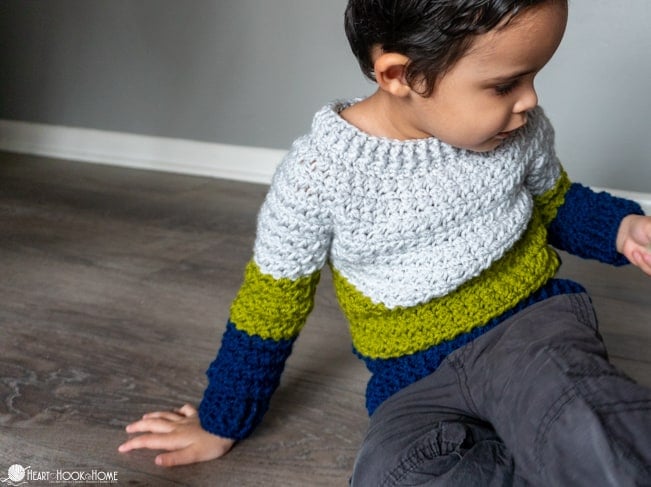 White, green, and blue colorway example of a child size Color Block Sweater crochet pattern on a male child wearing gray pants sitting on the floor.