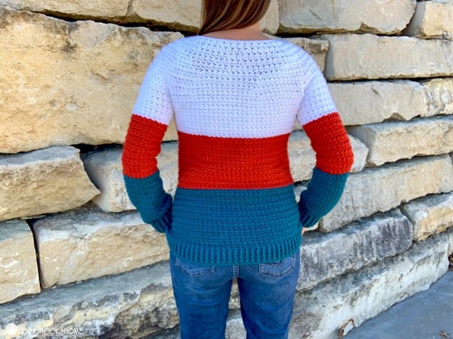 Back side of a White, Paprika, and Tidepool colored Color Block Sweater crochet pattern on a female model wearing jeans and standing in front of a brick wall.