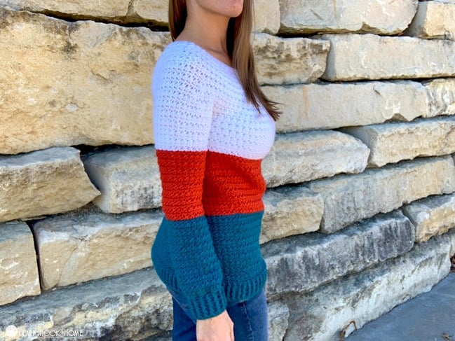 Side view of a White, Paprika, and Tidepool colored Color Block Sweater crochet pattern on a female model wearing jeans and standing in front of a brick wall.
