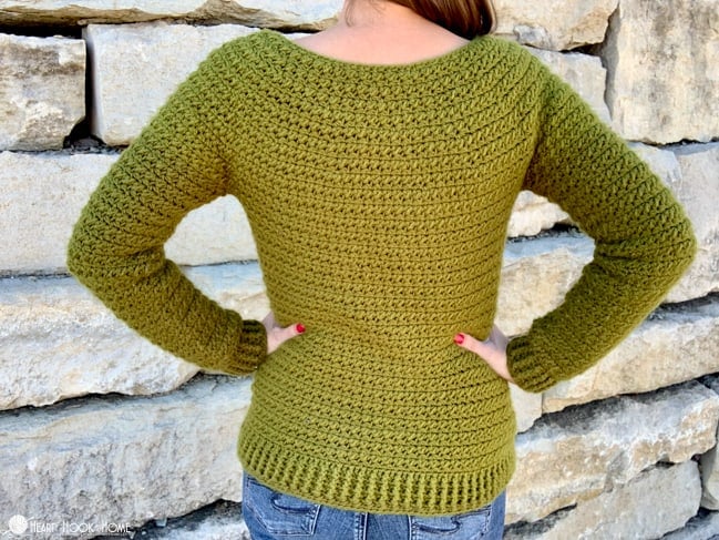 Back side of an olive colored Color Block Sweater crochet pattern on a female model wearing jeans and standing in front of a brick wall.