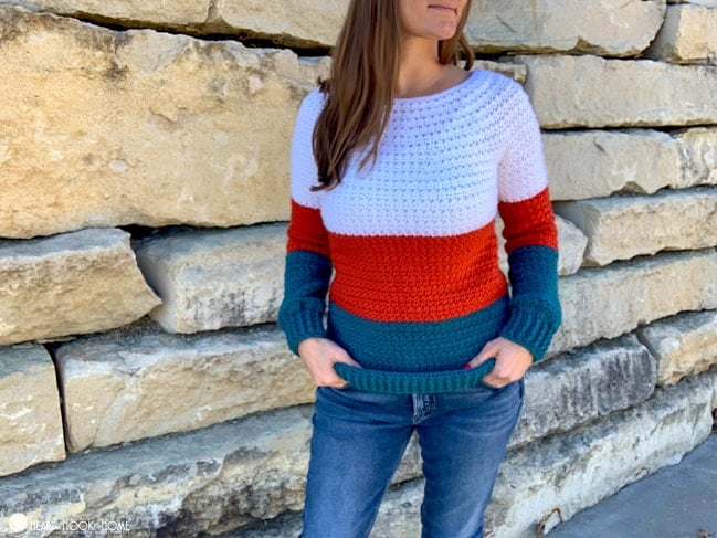 White, Paprika, and Tidepool colored Color Block Sweater crochet pattern on a female model wearing jeans and standing in front of a brick wall.