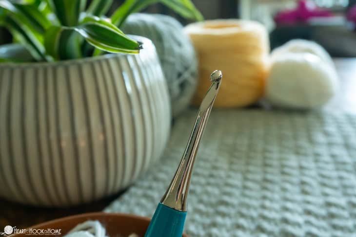 Blue Furls Odyssey crochet hook with skeins of yarn and a sample of the Crochet Stitch Sampler Afghan Pattern laying in the background.