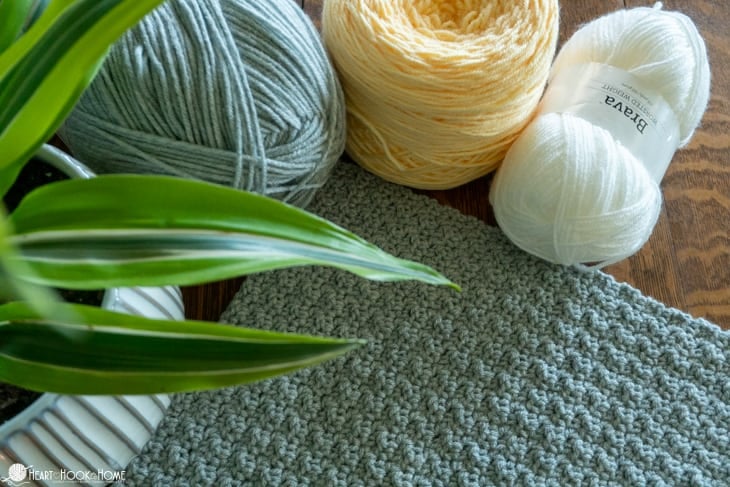Skeins of gray, yellow, and white yarn and a sample of the Crochet Stitch Sampler Afghan Pattern laying in front of them.