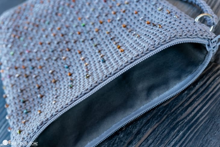 Gray sample of the Cross-Body Bead Bag crochet pattern with natural colored beads laying on its side showing the zipper and lining.