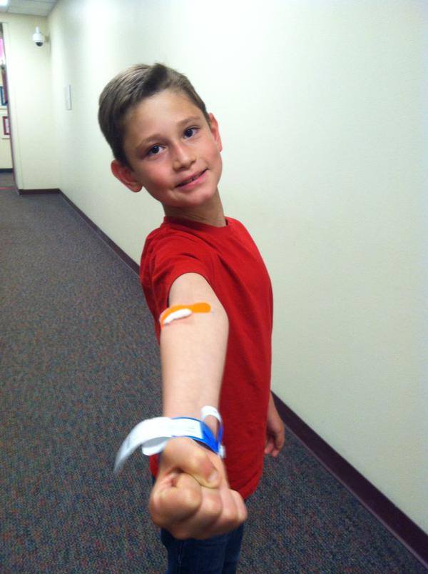 Smiling male child showing off a hospital bracelet and a Band-Aid covering a cotton ball. 