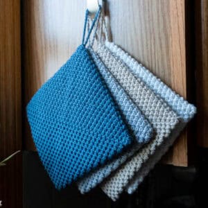 Gray, tan, and blue colorways of 8/6 Rainbow Cotton from Hobbii examples of the Double Thick Hot Pad crochet pattern hanging from a hook on a shelf.