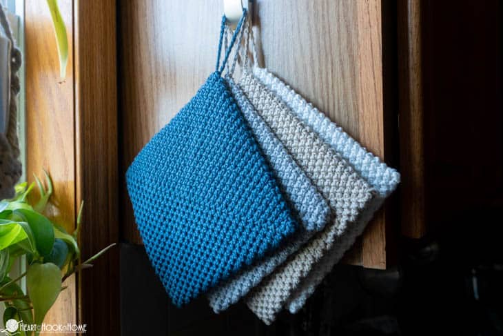 Gray, tan, and blue colorways of 8/6 Rainbow Cotton from Hobbii examples of the Double Thick Hot Pad crochet pattern hanging from a hook on a shelf.