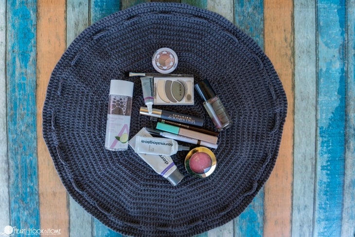 Example of a laid open, gray Drawstring Cosmetics Bag crochet pattern with various make up containers laying on it.