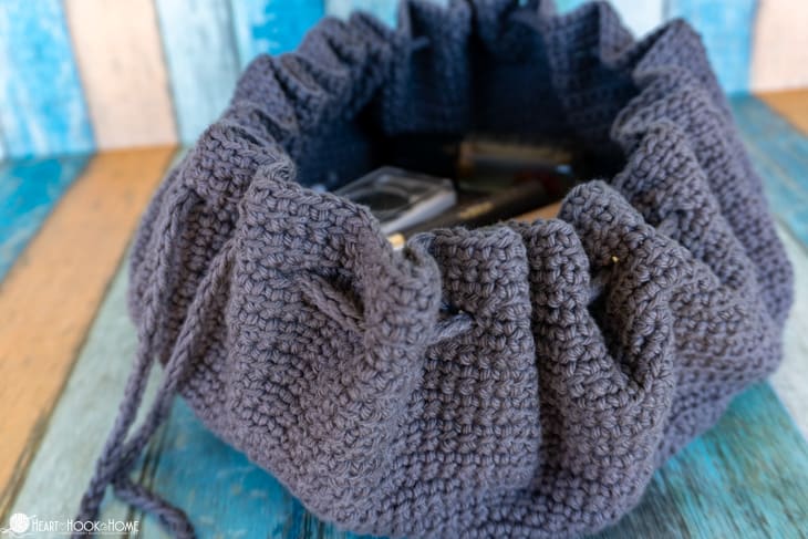 Example of a partially open, gray Drawstring Cosmetics Bag crochet pattern with various make up containers inside.