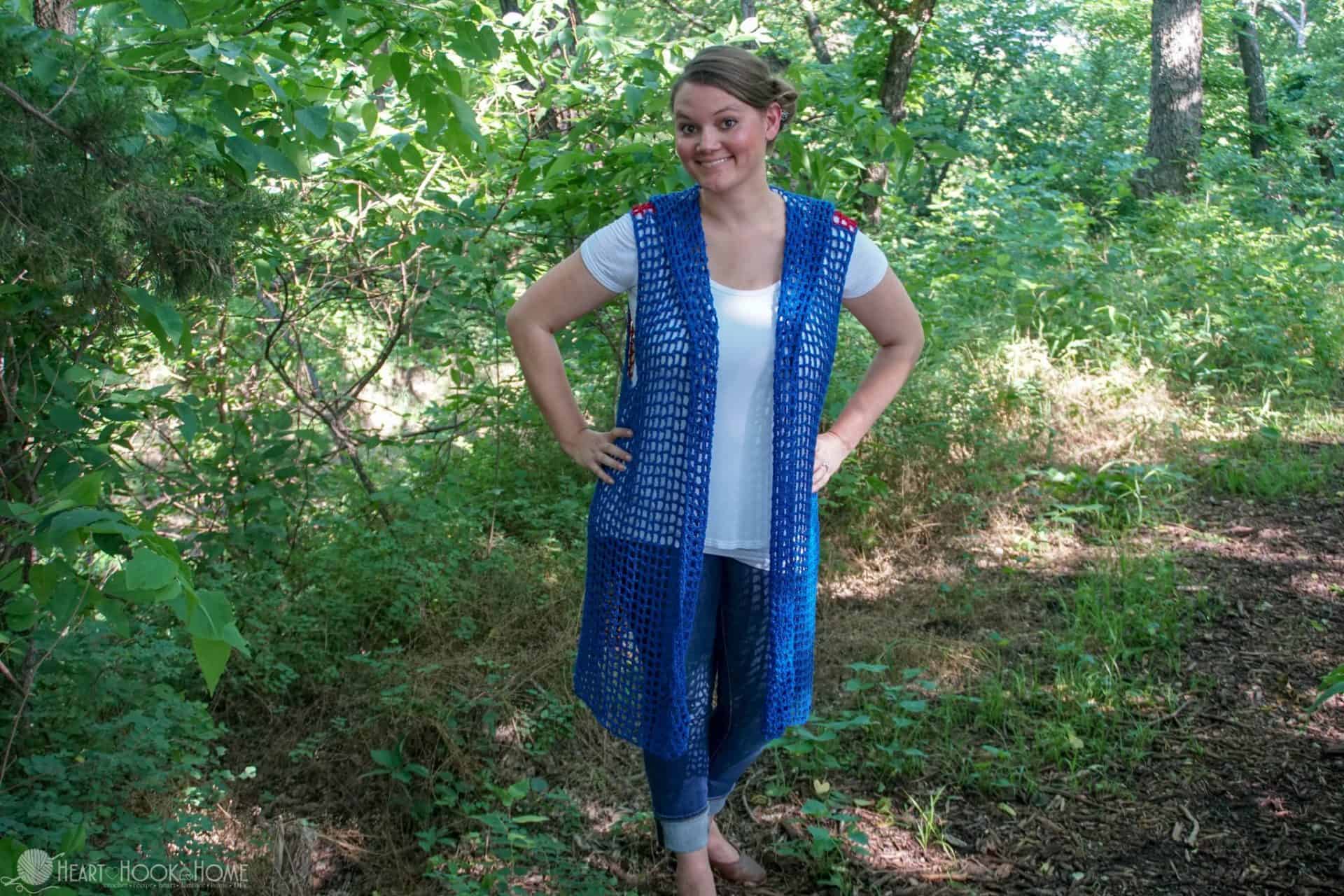Smiling female model wearing a blue example of the Sweet Summertime Lightweight Hooded Vest crochet pattern, a white shirt, and jeans in a shady forest setting.