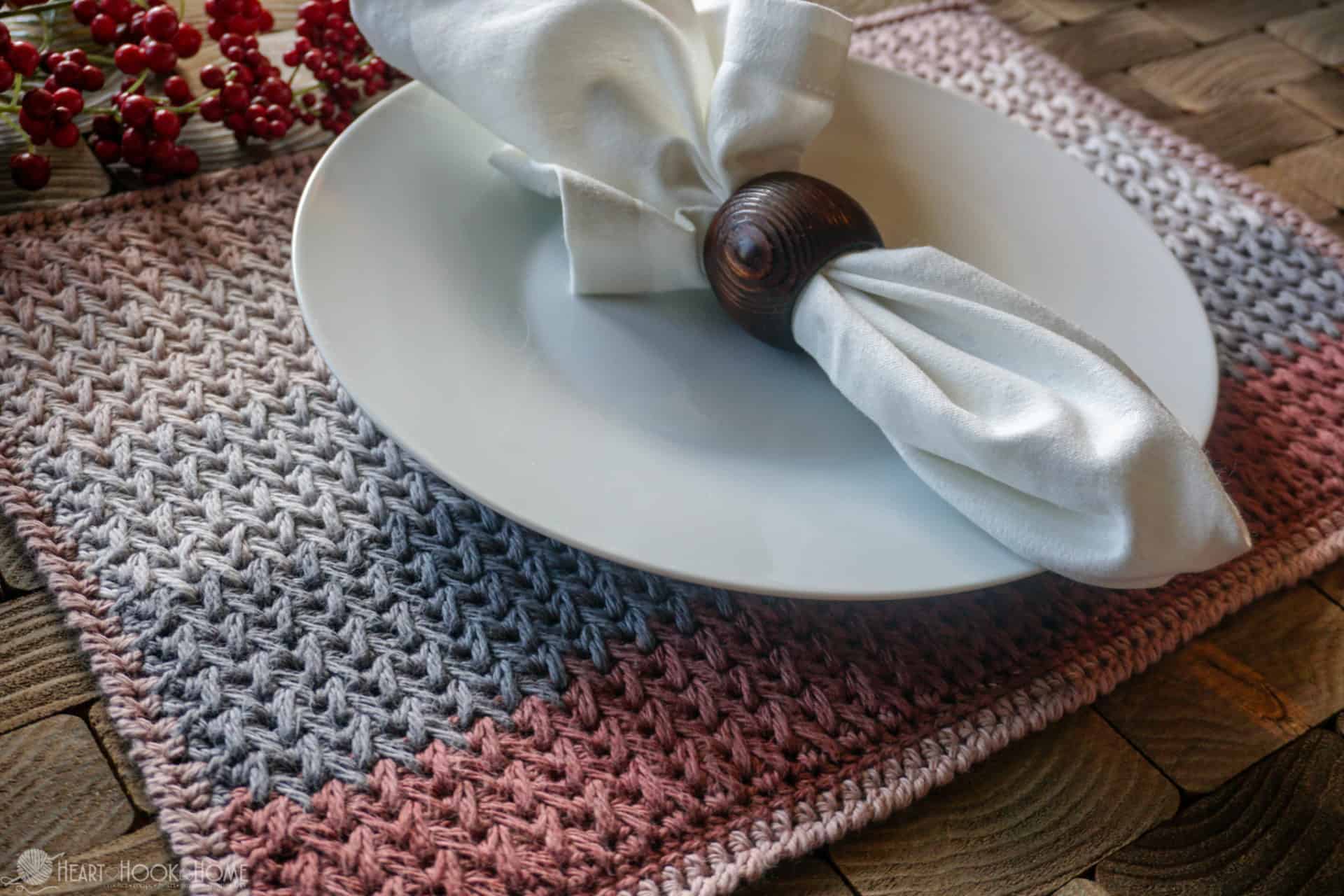 Example of a Nutmeg Nibbles crochet placemat pattern with a white plate and a white cloth napkin with a dark wood napkin ring sitting on it.