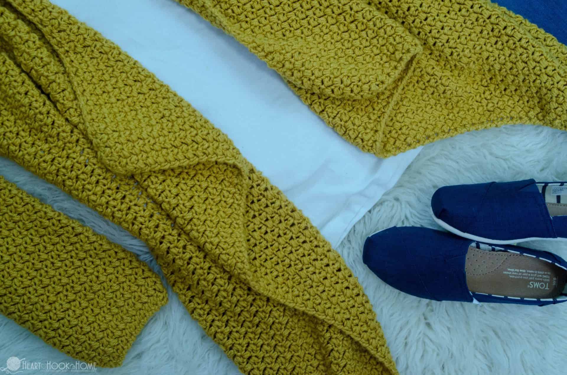 Gold example of The Elizabeth Cardigan crochet pattern over a white shirt and blue shoes nearby.