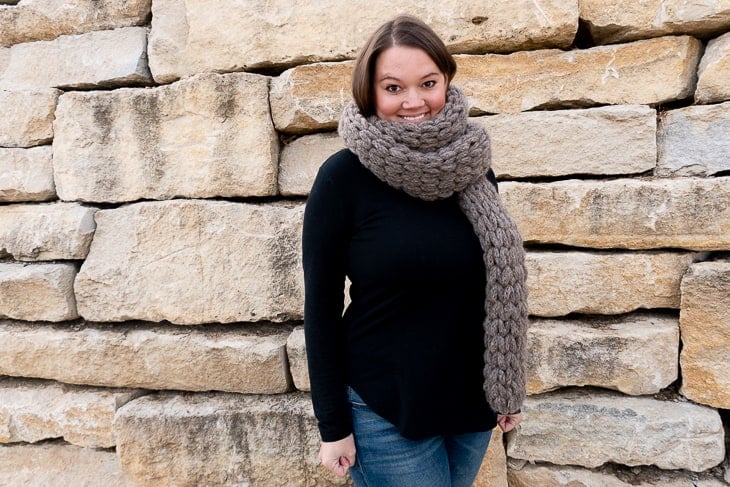 Smiling female model wearing a dark gray example of the Easy Loop Yarn Scarf and a black sweater and jeans while standing in front of a brick wall.