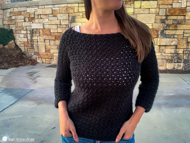 Female model wearing the Easy Peasy Pullover crochet pattern in an Asphalt Heather colorway of the Brava 500 yarn from WeCrochet standing in front of a brick wall.
