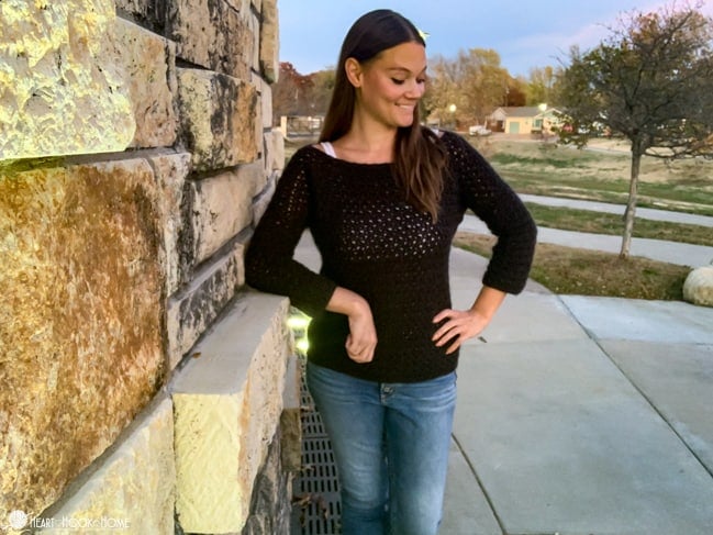 Smiling female model wearing the Easy Peasy Pullover crochet pattern in an Asphalt Heather colorway of the Brava 500 yarn from WeCrochet and jeans standing in front of a brick wall.