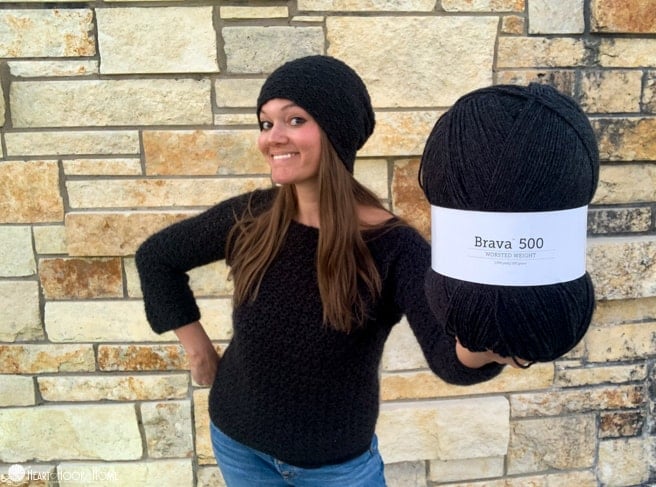 Smiling female model wearing an example of the Bricktown Slouch Beanie crochet pattern and the Easy Peasy Pullover crochet pattern both in an Asphalt Heather colorway of the Brava 500 yarn from WeCrochet and holding a skein of Brava 500 yarn in front of a brick wall.