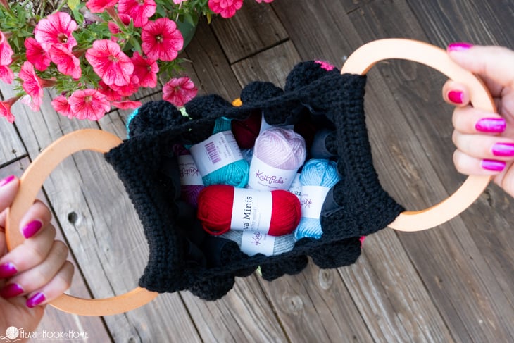 A 24-pack assortment of Brava Mini Skeins from WeCrochet in an example of a black colorway of the Fat Bottom Bag crochet pattern on a deck and near some pink flowers.
