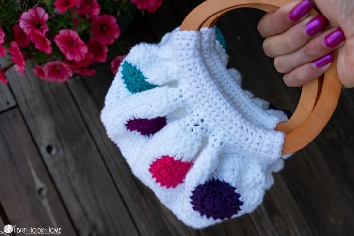 An example of a white, teal, purple, and pink colorway of the Fat Bottom Bag crochet pattern on a deck and near some pink flowers and being held in a hand.
