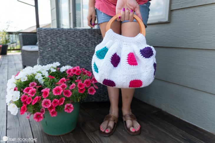 An example of a white, teal, purple, and pink colorway of the Fat Bottom Bag crochet pattern on a deck and near some pink flowers and being held by a female wearing jean shorts.