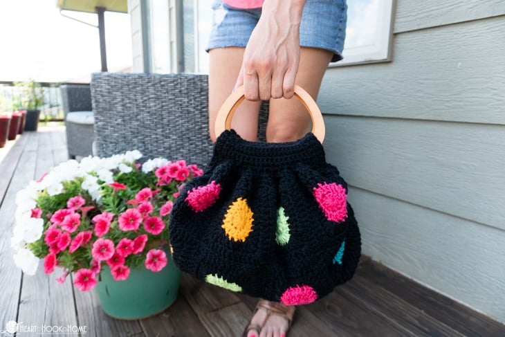 An example of a black colorway of the Fat Bottom Bag crochet pattern on a deck and near some pink flowers and being held by a female wearing jean shorts.