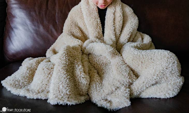 White example of the Faux Fur Throw crochet pattern draped around a teen model sitting on a leather couch.