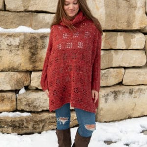 Smiling female model wearing jeans, boots and a rust colored example of the Filet Poncho crochet pattern standing in the snow in front of a stone wall.
