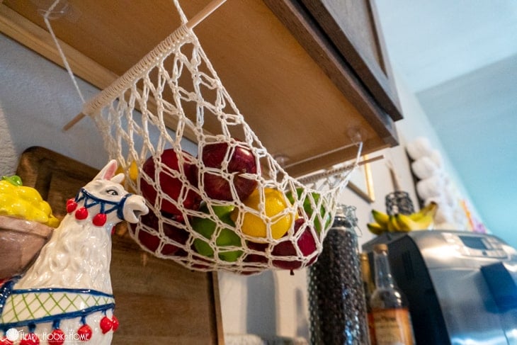 Example of a Fruit Hammock crochet pattern in a white colorway with apples, oranges, and lemons nestled in it. The hammock is attached to the underside of a kitchen cabinet.