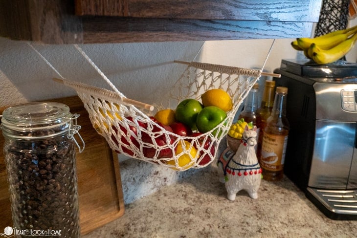 Example of a Fruit Hammock crochet pattern in a white colorway with apples, oranges, and lemons nestled in it. The hammock is hanging from the underside of a kitchen cabinet.