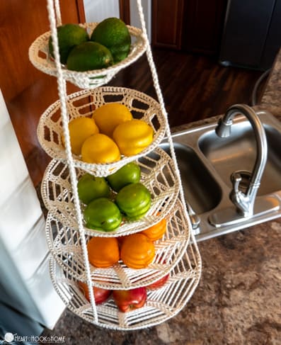 White colorway example of the Hanging Baskets crochet pattern hanging in a kitchen setting near a sink. Each basket has with avocadoes, lemons, limes, oranges, and apples sitting in it.