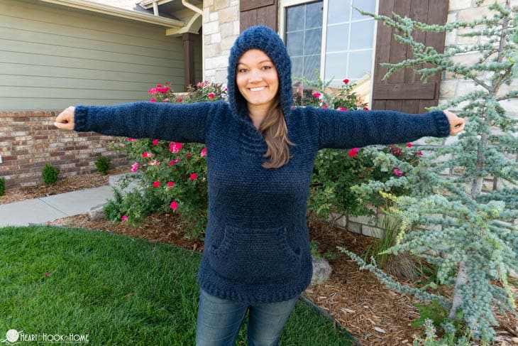 Blue example of the Hibernation Hoodie crochet pattern on a smiling female model standing with her arms outstretched in a front yard setting.
