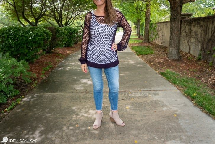 Black colorway of the Hot Mesh Summer crochet pattern on a smiling female model wearing a white tank top underneath it and jeans in a park setting. 