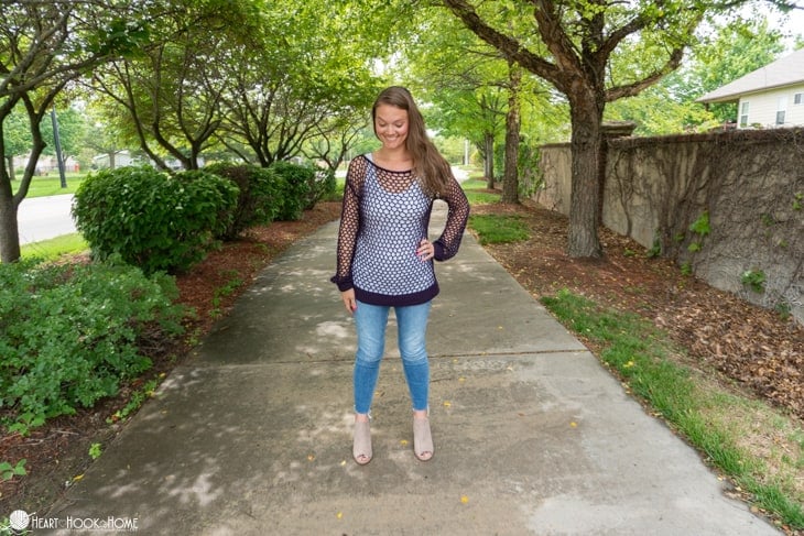 Black colorway of the Hot Mesh Summer crochet pattern on a smiling female model wearing a white tank top underneath it and jeans in a park setting.  