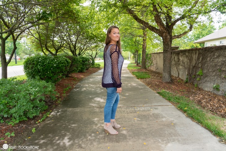 Side view of a black colorway of the Hot Mesh Summer crochet pattern on a smiling female model wearing a white tank top underneath it and jeans in a park setting. 