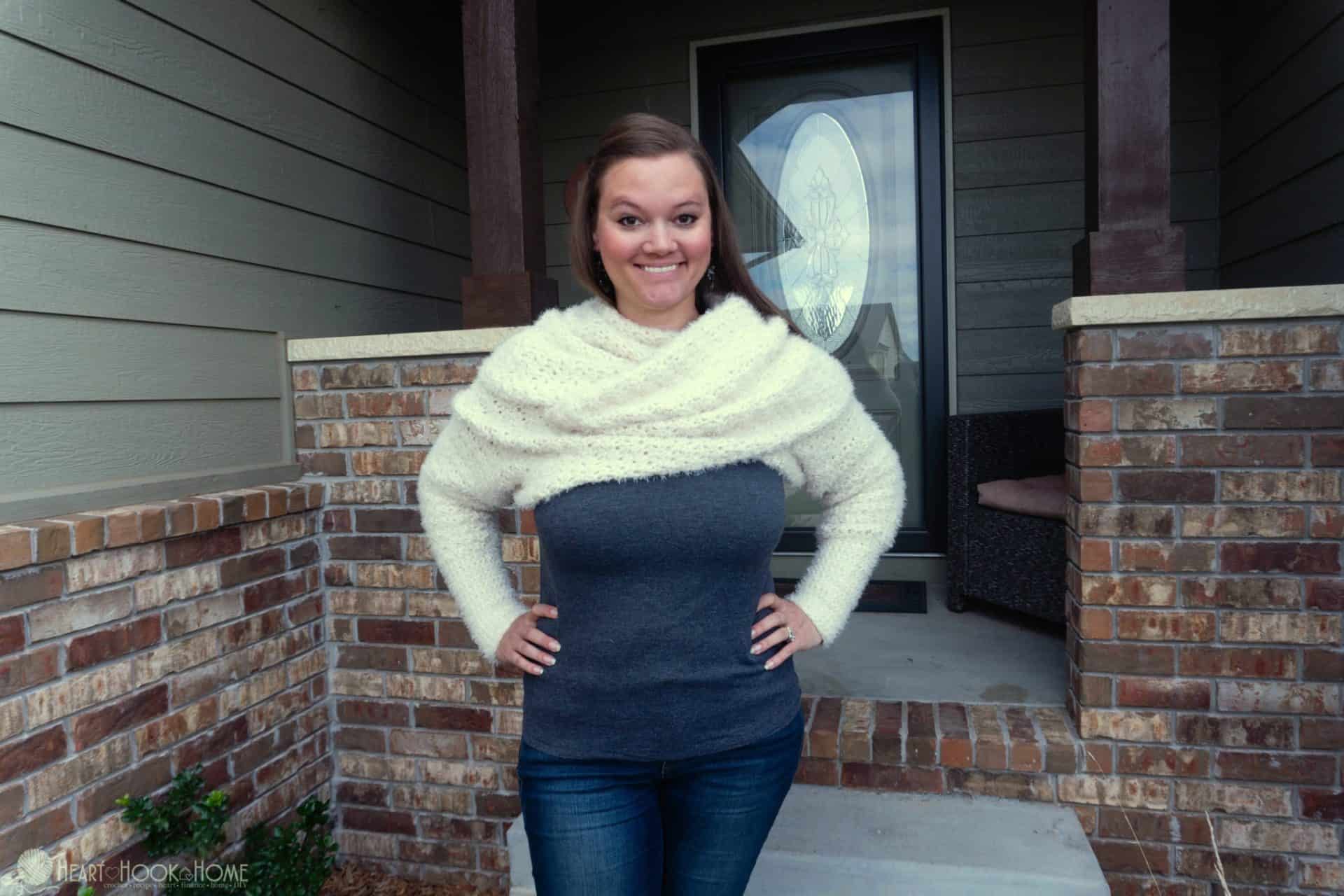 White example of the Give Me A Hygge Scarf with Sleeves crochet pattern on a smiling female model wearing a gray shirt and jeans and standing in a front door setting.