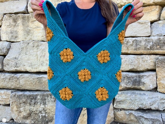 Blue and gold example of the Marigold Sling Bag crochet pattern being held by a female in front of a brick wall. 