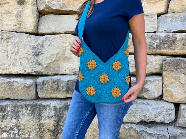 Female model wearing a blue t-shirt and jeans and standing in front of a brick wall. She is modeling an example of the Marigold Sling Bag crochet pattern in a Mustard Seed and Surf colorway of Cotlin Yarn.