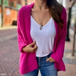 Example of the Odette Cardigan Tunisian Crochet Pattern in a bright pink on a female model with a white undershirt and jeans on a cobblestone street.
