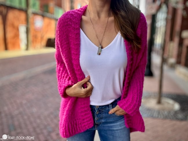 Example of the Odette Cardigan Tunisian Crochet Pattern in a bright pink on a female model with a white undershirt and jeans on a cobblestone street.
