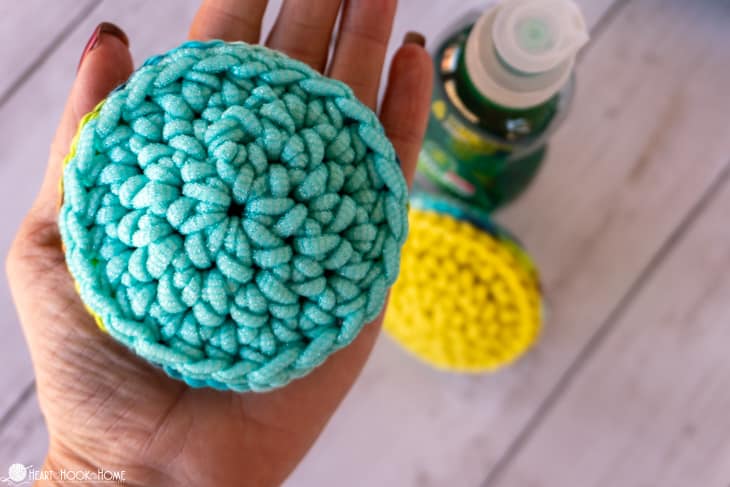 Example of the Pot Scrubber crochet pattern in a teal colorway being held in a hand with a yellow colorway next to a bottle of dish soap in the background.