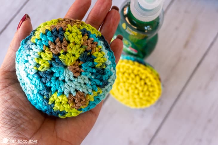 Example of the Pot Scrubber crochet pattern in a variegated colorway being held in a hand with a yellow colorway next to a bottle of dish soap in the background.