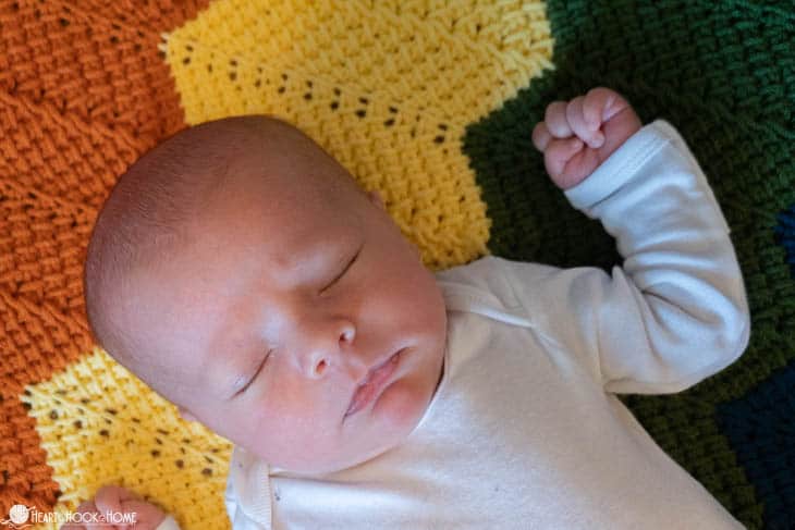Baby in a white onesie asleep on a rainbow colored example of the Rainbow Baby Blanket crochet pattern.