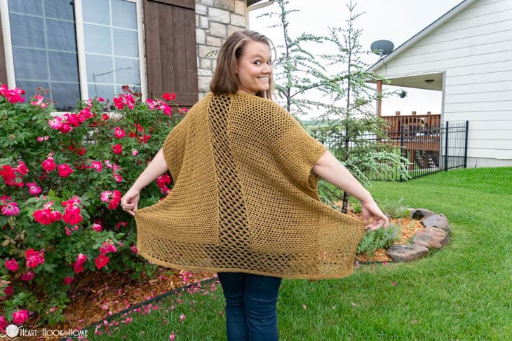 Smiling female model showing off the back of a Yarn Bee Soft & Sleek DK in “Dijon” colorway example of the Come On I Wanna Ruana crochet pattern in a front yard setting.