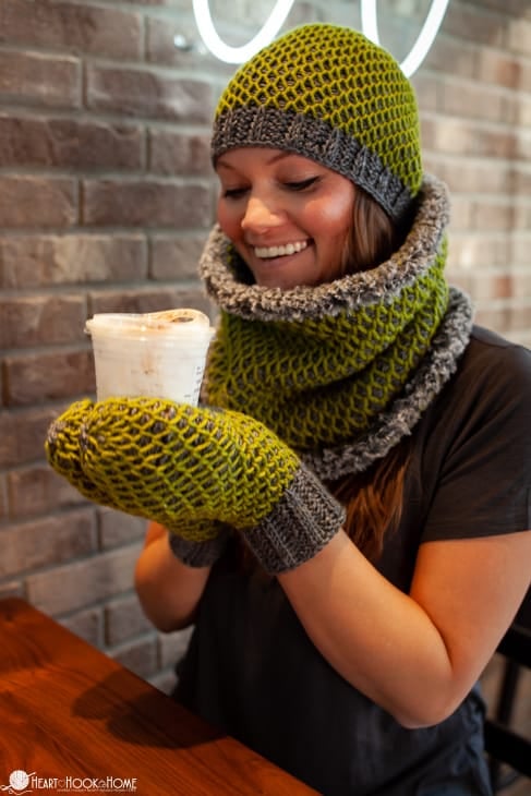 Tunisian “Smock it to Me” Crochet Set in Avocado and Cobblestone Heather colorways of Brava worsted and bulky yarn on a smiling female model holding a cup of coffee by a brick wall.